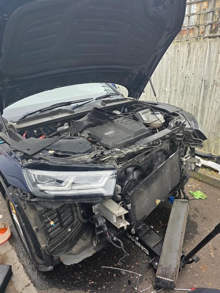 Audi with its front bumper off and radiator pulled out, parked by a garden fence