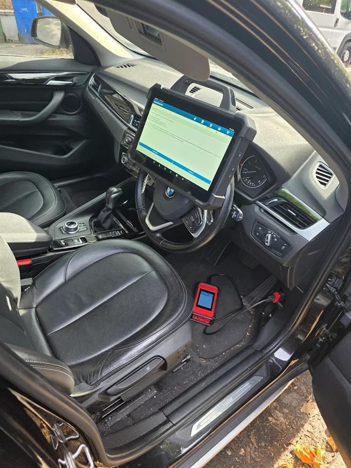 Diagnostic tablet on a BMW X1 steering wheel with a battery tester clipped in on the floor